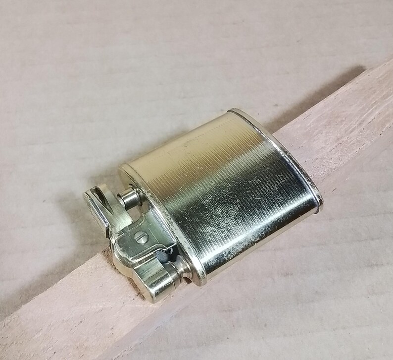 May include: A vintage, metallic gold-colored lighter with a textured surface. The lighter is rectangular with rounded edges and a hinged top. It rests on a light-colored wooden stick, showcasing its design and details.