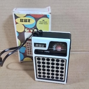 Könnte beinhalten: Ein Vintage EEI AM Solid-State-Taschenradio mit weißem und schwarzem Gehäuse. Das Radio hat ein Zifferblatt, Lautsprecherlöcher und eine Handschlaufe. Die Box zeigt das EEI-Logo und bunte Kreise.