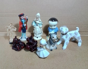 Puede incluir: Una colección de figuritas de porcelana vintage, que incluye una mujer con un vestido blanco, un hombre con un abrigo rojo, un niño con una camisa azul, una niña con un vestido blanco, un perro y dos figuritas pequeñas.