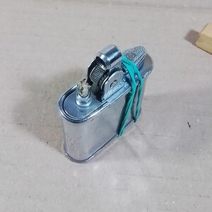May include: A vintage silver-colored lighter with a textured top and a green rubber band wrapped around it. The lighter has a rectangular body and a small, cylindrical nozzle. A small wooden block is in the background.
