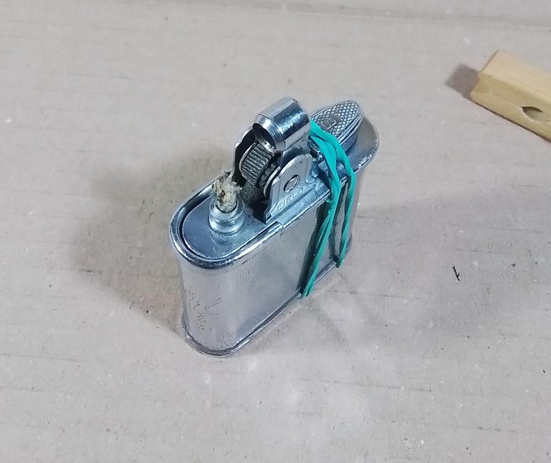May include: A vintage silver-colored lighter with a textured top and a green rubber band wrapped around it. The lighter has a rectangular body and a small, cylindrical nozzle. A small wooden block is in the background.
