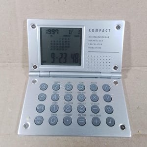 May include: A silver compact digital calendar with a black LCD screen displaying the date 9-23-48. The calendar also has a calculator, alarm clock, and world time functions. The buttons are labeled with city names and functions.