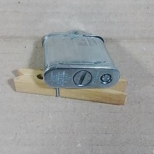 May include: A vintage silver-colored metal lighter with a textured surface. The lighter is rectangular with rounded edges and a hinged top. It rests on a wooden clothespin. The lighter has text on the bottom.