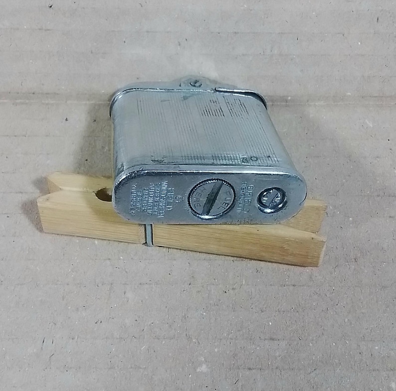 May include: A vintage silver-colored metal lighter with a textured surface. The lighter is rectangular with rounded edges and a hinged top. It rests on a wooden clothespin. The lighter has text on the bottom.
