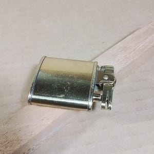 May include: A vintage gold-toned metal lighter with a rectangular body and a hinged top. The lighter has a textured surface and a detailed ignition mechanism. It rests on a light-colored wooden surface.