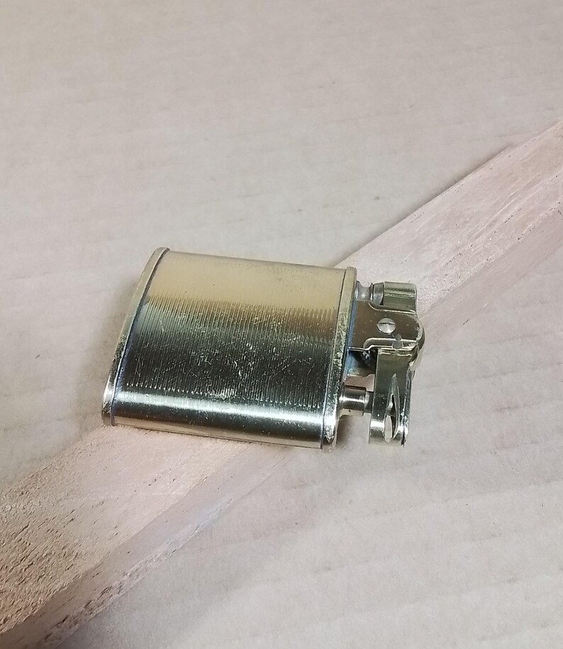 May include: A vintage gold-toned metal lighter with a rectangular body and a hinged top. The lighter has a textured surface and a detailed ignition mechanism. It rests on a light-colored wooden surface.