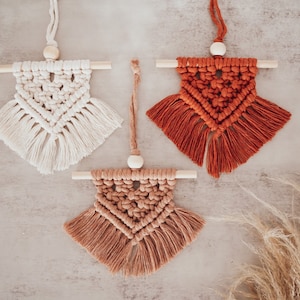 May include: Three macrame wall hangings with wooden dowels and fringe. The hangings are in shades of white, brown, and orange.