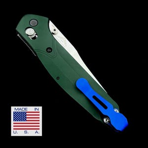 May include: A green pocket knife with a blue pocket clip. The knife has a partially opened blade and a silver handle. The knife is made in the USA.