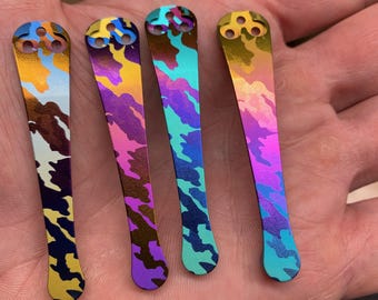 May include: Four decorative metal knife scales with a camouflage design in blue, yellow, purple, and green. Each scale features three circular holes at the top. The scales are held in a hand.