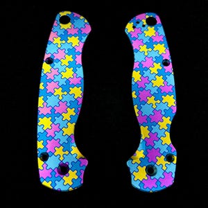 May include: Two knife scales with a colourful jigsaw puzzle pattern. The scales are in shades of blue, yellow, and pink. The scales have holes for screws and a curved shape. The background is black.