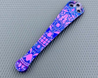 May include: A blue and pink titanium pocket tool with a tiki-inspired design. The tool has a textured surface and a hole at the top.