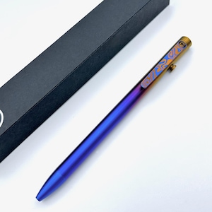 May include: A sleek, metallic pen with a gradient finish, transitioning from blue to purple. The pen's top section features a gold-toned accent with an orange and blue patterned design. A black pen box is in the background.