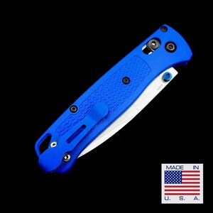 May include: A vibrant blue folding knife with a textured handle and a silver blade. The knife features a pocket clip and is set against a black background. The image includes a small "Made in U.S.A." sticker.