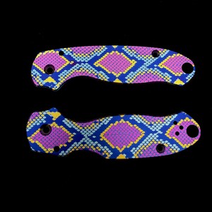 K&ouml;nnte beinhalten: Zwei Messergiffschalen mit einem lebendigen Schlangenhautmuster. Das Design zeigt ein sich wiederholendes Muster aus rosa, gelben und hellblauen Rauten auf einem dunkelblauen Hintergrund. Die Schalen haben L&ouml;cher f&uuml;r Schrauben und eine gebogene Form.
