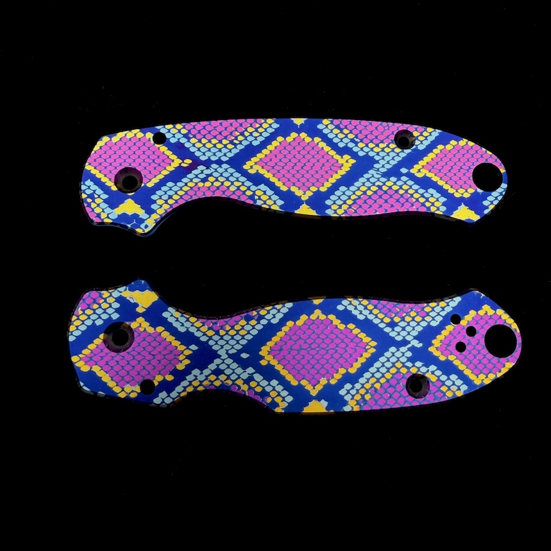 K&ouml;nnte beinhalten: Zwei Messergiffschalen mit einem lebendigen Schlangenhautmuster. Das Design zeigt ein sich wiederholendes Muster aus rosa, gelben und hellblauen Rauten auf einem dunkelblauen Hintergrund. Die Schalen haben L&ouml;cher f&uuml;r Schrauben und eine gebogene Form.