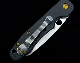 May include: A black and silver folding knife with a carbon fiber handle. The knife features a silver pocket clip with a spider logo and gold-colored screws. The blade is partially open, revealing a silver edge.