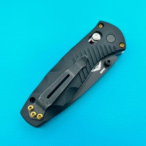 May include: A black folding knife with a partially serrated blade. The handle features a camouflage pattern and a pocket clip. Gold-colored screws accent the handle. The blade has the text "18401" printed on it.