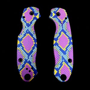 K&ouml;nnte beinhalten: Zwei Messerschaftschalen mit einem lebendigen Schlangenhautmuster. Die Schalen weisen ein sich wiederholendes Design aus rosa, blauen und gelben Rauten auf dunklem Hintergrund auf. Jede Schale hat mehrere L&ouml;cher zur Befestigung.
