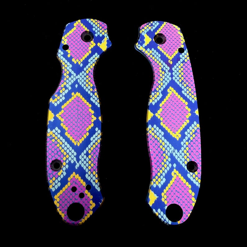 K&ouml;nnte beinhalten: Zwei Messerschaftschalen mit einem lebendigen Schlangenhautmuster. Die Schalen weisen ein sich wiederholendes Design aus rosa, blauen und gelben Rauten auf dunklem Hintergrund auf. Jede Schale hat mehrere L&ouml;cher zur Befestigung.