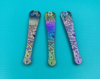 May include: Three decorative metal pieces with detailed geometric patterns in gold, purple, and blue hues. The elongated, curved pieces have a hole at the top. They are displayed on a turquoise background, showcasing their vibrant colours.