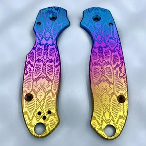 May include: Two knife scales with a vibrant, iridescent finish. The scales feature a snakeskin pattern in a gradient of colors, transitioning from blue to purple, then to yellow. Each scale has pre-drilled holes for attachment.