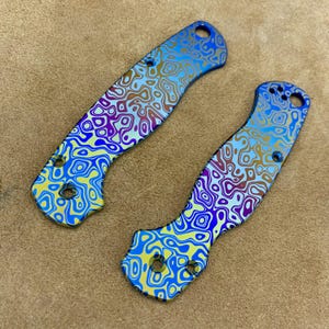 Può includere: Due guancette per manico di coltello con un vivace motivo a vortice nelle tonalità del blu, giallo, viola e marrone. Le guancette hanno fori per il fissaggio e un design ergonomico curvo. Lo sfondo è una superficie marrone.