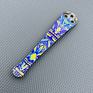 Puede incluir: Un cuchillo de bolsillo de titanio con un diseño único de patrón azul y amarillo. El cuchillo tiene una hoja pequeña y redondeada y un mango texturizado.
