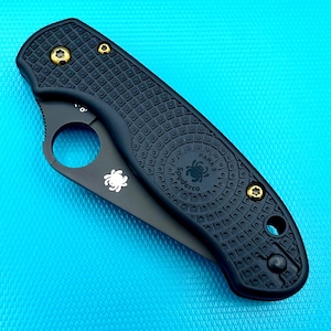 May include: Black folding knife with a textured handle and a partially serrated blade. The knife features a circular opening for one-handed use and gold-colored screws. The blade has a white spider logo and the text "Spyderco PARA 3".