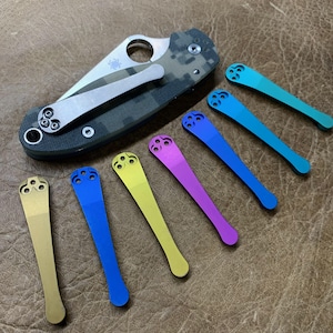 May include: A folding pocket knife with a camouflage handle and a silver blade, next to an assortment of colorful pocket clips. The clips are in shades of gold, blue, yellow, purple, and teal.