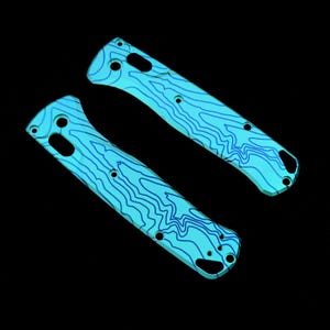 Puede incluir: Dos cachas de cuchillo turquesas con un diseño de mapa topográfico en azul oscuro. Las cachas tienen múltiples agujeros para tornillos y una forma curva. El fondo es negro.