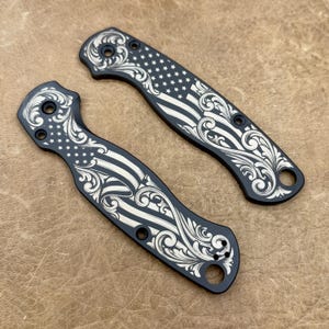 May include: Two black knife scales with engraved American flag and scrollwork designs. The scales feature the stars and stripes of the flag, along with decorative flourishes. The scales have holes for attachment and a smooth, matte finish.