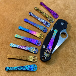 May include: A set of ten colourful titanium scales with a swirling pattern. The scales are arranged in a row, with a black pocket knife at the end of the row.