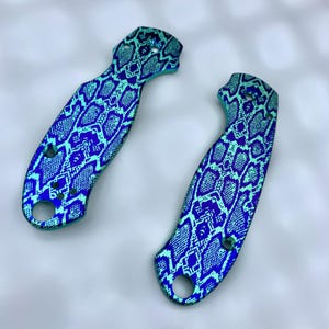 Op de afbeelding: Twee mesgrepen met een blauw en teal slangenpatroon. De grepen hebben een gebogen vorm met gaten voor schroeven. De kleurovergang verschuift van teal naar blauw, waardoor een opvallend visueel effect ontstaat. De grepen zijn ontworpen voor een mes.