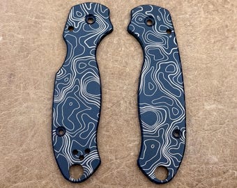 May include: Two dark blue knife handle scales with a white topographic pattern. Each scale has a curved shape with holes for screws and a lanyard hole. The scales are made of metal and are designed to replace existing knife handles.