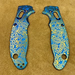 Puede incluir: Dos cachas de cuchillo con un patrón arremolinado azul y dorado. Las cachas tienen una forma curva con múltiples agujeros para tornillos y un agujero circular en la base. El diseño recuerda al agua que fluye o un mapa topográfico.