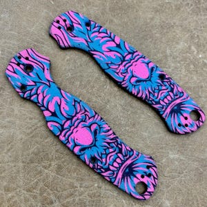 May include: Two knife handle scales with a vibrant pink and blue floral design. The scales are curved and feature holes for attachment. The design includes a central pink shape surrounded by blue and pink floral patterns.