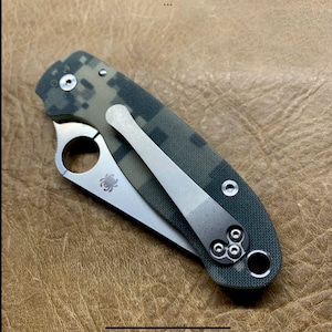 May include: A green and brown camouflage pocket knife with a silver blade and a silver pocket clip. The knife has a spider logo on the blade.