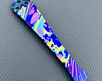 May include: A blue titanium pocket tool with a colourful abstract pattern in pink, yellow, and blue. The tool has a rounded tip and a hole at the top.