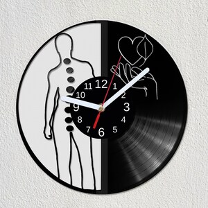 Puede incluir: Un reloj de pared en blanco y negro hecho de un disco de vinilo. La esfera del reloj presenta una silueta humana y una mano que sostiene un corazón y una hoja. Las manecillas del reloj son blancas con un segundero rojo.