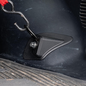 Puede incluir: Accesorio de coche de plástico negro con gancho y perno metálico. El accesorio está montado sobre una superficie oscura, posiblemente el interior de un vehículo. Una correa roja está sujeta al gancho.