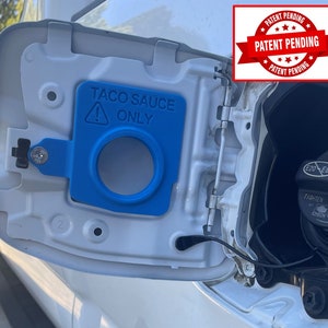 May include: A white car gas tank door with a blue plastic insert that says "Taco Sauce Only" with an exclamation point. The door is open and the gas cap is visible. A red sticker on the car says "Patent Pending" with stars around the text.
