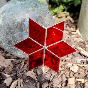 Könnte beinhalten: Ein roter Glasstern-Anhänger mit einem silbernen Drahtbügel. Der Stern besteht aus vielen kleinen Glasscheiben, die mit Blei zusammengefügt sind. Der Stern hängt an einem Ast.