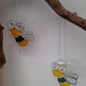 Könnte beinhalten: Zwei gelbe und schwarze Glas-Bienen-Ornamente, die an einem Ast hängen. Die Bienen haben weiße Augen und sind an silbernen Schnüren aufgehängt.