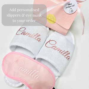 May include: A white spa set featuring personalized slippers and an eye mask, all with the name "Camilla" in a rose gold script. The set includes a pink gift box labeled "Mini pamper set".