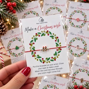 May include: A red string bracelet featuring a small gift-shaped charm on a white display card. The card displays a green wreath illustration, the text Make a Christmas wish, and instructions to close your eyes and make a wish.