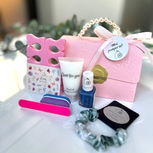 May include: A pink gift box with a pearl handle, containing a mini pamper set. The set includes a nail file, nail polish, hand lotion, toe separators, nail stickers, and a scrunchie. The nail polish is a deep blue color.
