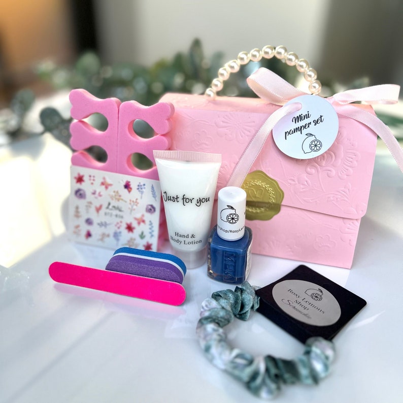May include: A pink gift box with a pearl handle, containing a mini pamper set. The set includes a nail file, nail polish, hand lotion, toe separators, nail stickers, and a scrunchie. The nail polish is a deep blue color.
