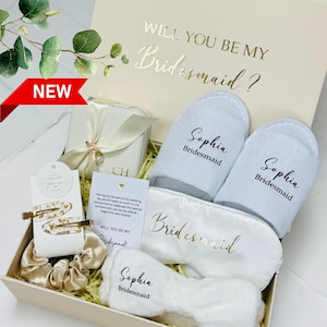 May include: A bridesmaid gift box containing light blue slippers, a white sleep mask, and a headband, all personalised with the name "Sophia". The box also includes hair accessories and a card with the text "Will you be my Bridesmaid?" in gold lettering.