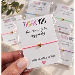 May include: A pink string bracelet featuring a small gold-coloured heart charm, presented on a white card inside a clear plastic sleeve. The card displays the text "THANK YOU for coming to my party" and "I hope you had a great time!" with a pink heart icon.
