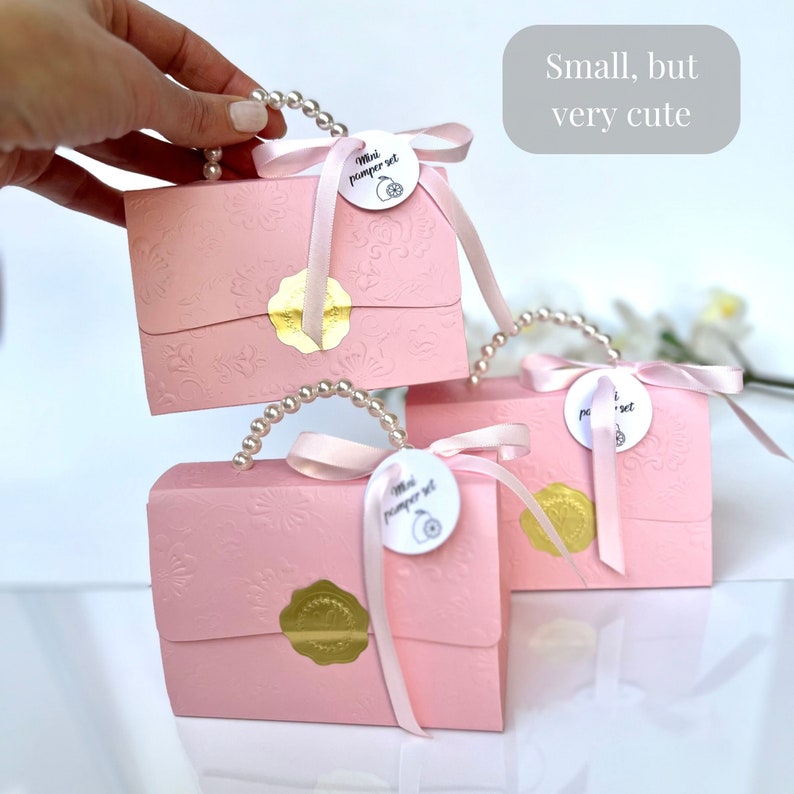 May include: Three pink gift boxes with embossed floral patterns, pearl handles, and pink ribbons. Each box has a gold seal and a tag that reads "Mini pamper set". The text "Small, but very cute" is in a gray rectangle.
